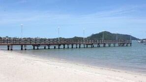 A beach with a long pier extending into the water, supported by concrete pillars, and a small hill with trees in the background. - Casa MonteFlor -Desacelere com gentileza (Governador Celso Ramos)