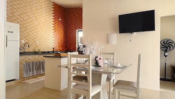 Un comedor con mesa de vidrio y sillas blancas, una cocina con paredes de ladrillo y un televisor de pantalla plana montado en la pared.