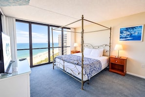 Una cama con dosel con vista a la playa, un escritorio con computadora y una mesita de noche con lámpara.