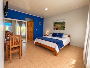 A bedroom with a bed, a desk with a chair, a window with curtains, and a painting on the wall. - Hotel Sueño de Luna (Aguas Zarcas)