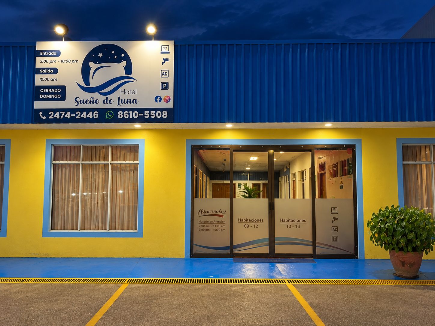 A hotel entrance with a blue and yellow facade, a reception desk, and a signboard with operating hours and contact information.