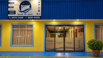 A hotel entrance with a blue and yellow facade, a reception desk, and a signboard with operating hours and contact information.