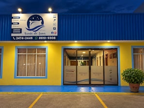 A hotel entrance with a blue and yellow facade, a reception desk, and a signboard with operating hours and contact information.