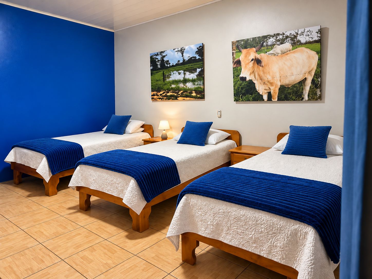 Une chambre d’hôtel avec trois lits simples, chacun recouvert d’une couverture bleue et doté de draps blancs. Deux tableaux sont accrochés au mur, l’un représentant un paysage et l’autre une vache.