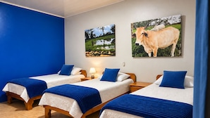 Ein Hotelzimmer mit drei Einzelbetten, jedes mit einer blauen Decke und weißer Bettwäsche. An den Wänden hängen zwei Bilder: eines zeigt eine Landschaft, das andere eine Kuh.