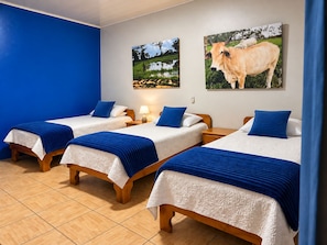 A hotel room with three single beds, each with a blue blanket and white bedding. There are two wall-mounted paintings, one of a landscape and one of a cow. - Hotel Sueño de Luna (Aguas Zarcas)