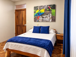 A bedroom with a bed, a nightstand, a wooden door, and a painting of colorful birds.