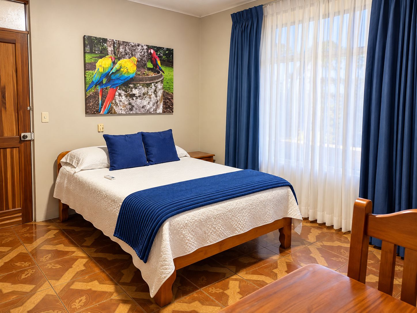 A hotel room with a bed, a nightstand, a chair, a painting of parrots, and a window with curtains.