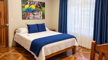 A hotel room with a bed, a nightstand, a chair, a painting of parrots, and a window with curtains.