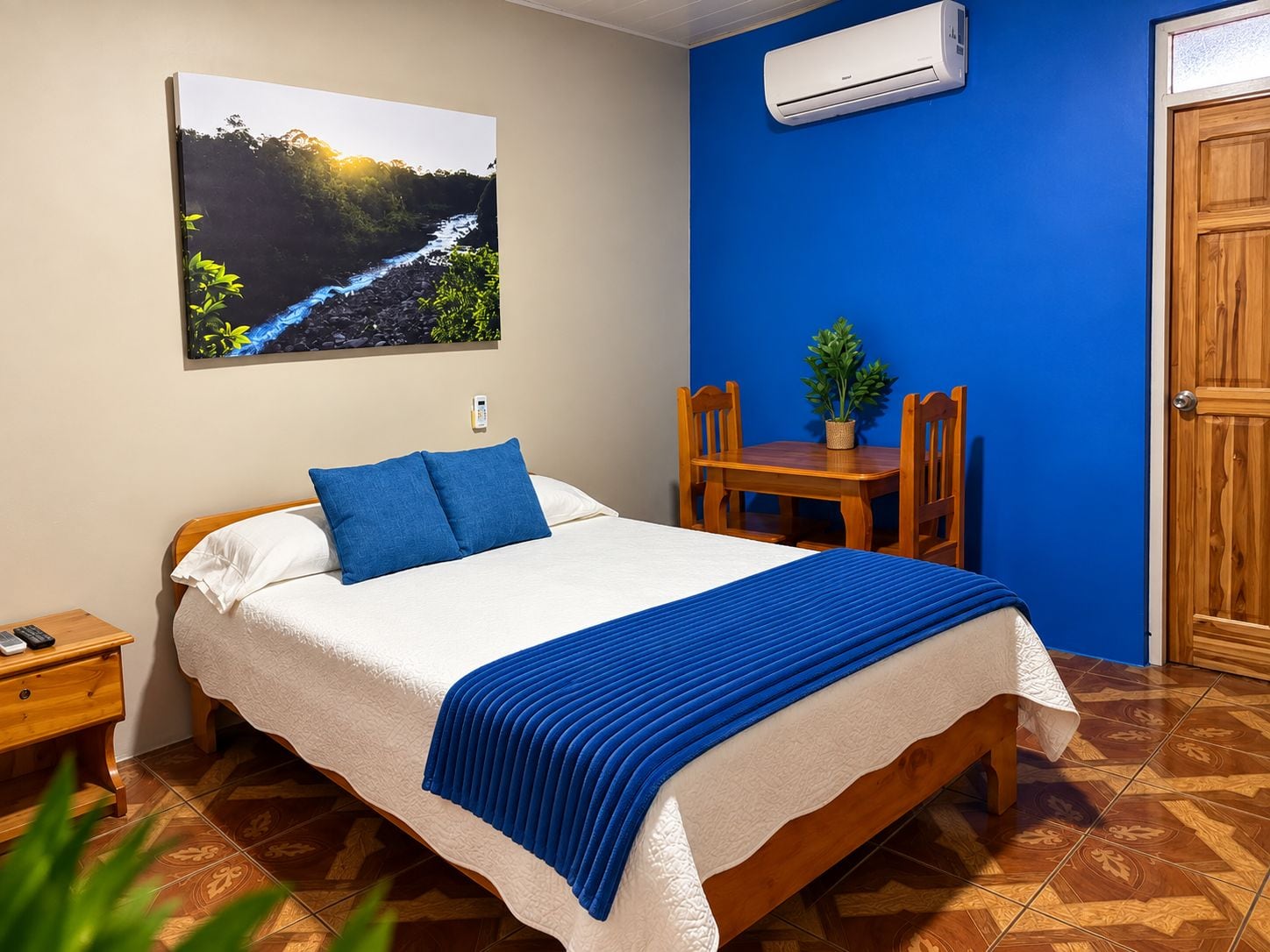 A bedroom with a bed, a nightstand, a table with chairs, a potted plant, and an air conditioning unit.