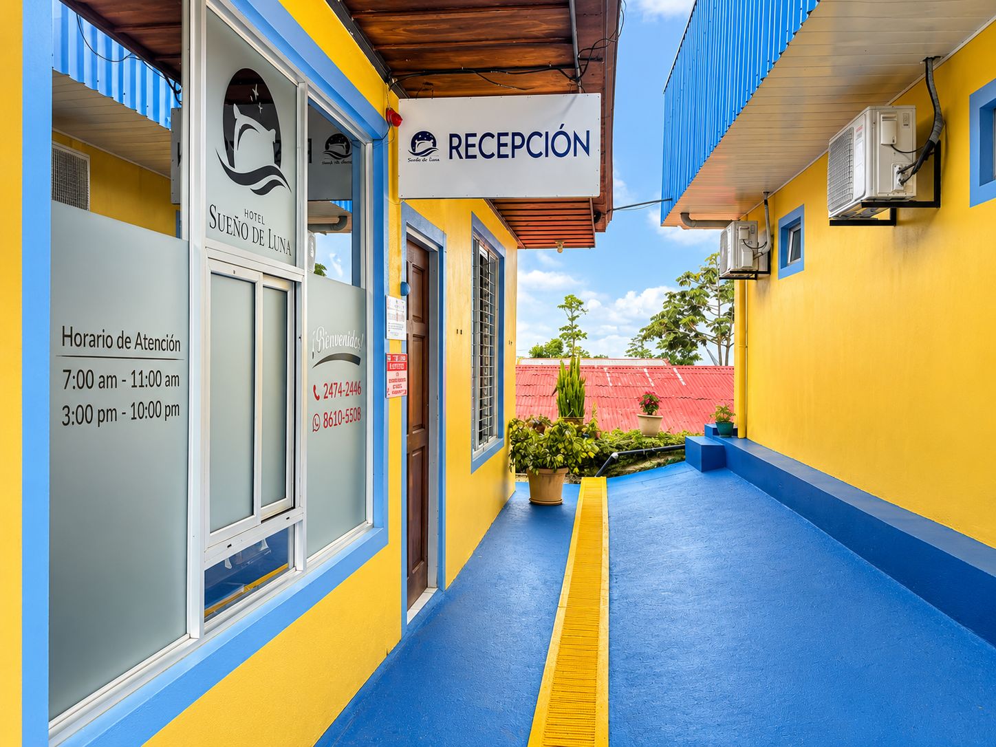 Zona de recepción de un hotel con mostrador, ventanal con vista al cielo y cartel con el nombre del hotel y horario de atención.