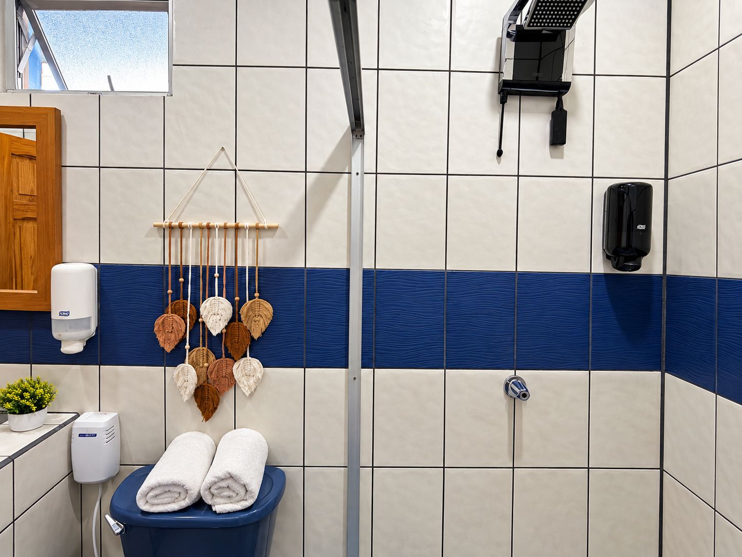 A bathroom with a blue and white tiled wall, a shower, a toilet with two towels, a soap dispenser, and a hanging decorative item.