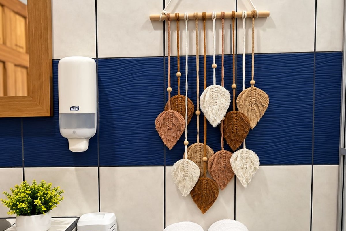 Ein Badezimmer mit einer blauen Toilette, einer hängenden Makramee-Dekoration, einem Handtrockner und einem Mülleimer.