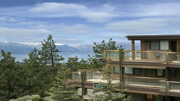 Un edificio a più piani con una targa per la reception, circondato da alberi e con vista su montagne e un lago.