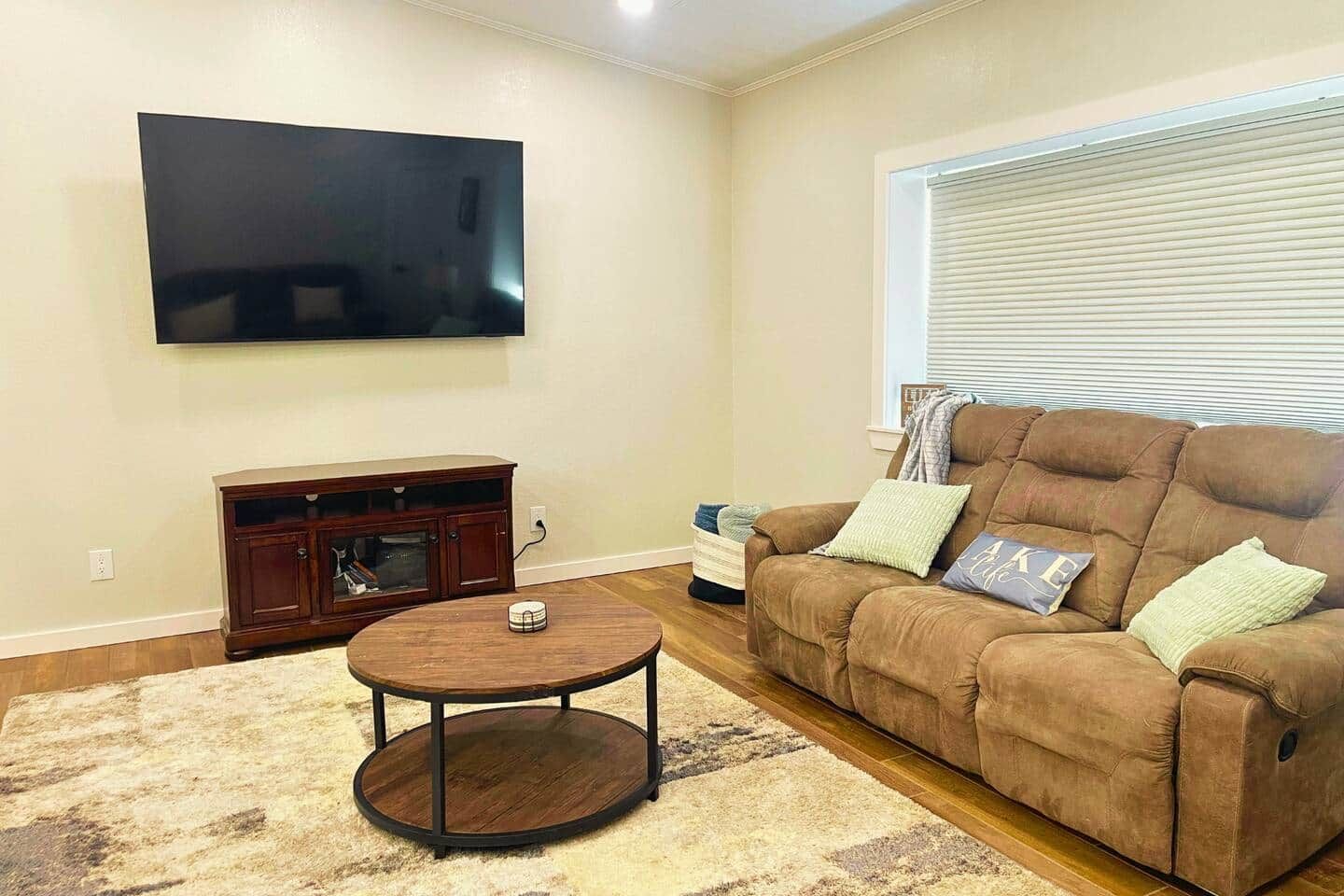 A living room with a flat-screen TV mounted on the wall, a brown leather sofa with cushions, a small wooden coffee table, and a patterned rug.