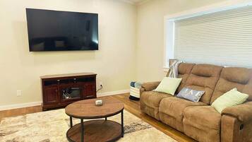 A living room with a flat-screen TV mounted on the wall, a brown leather sofa with cushions, a small wooden coffee table, and a patterned rug.