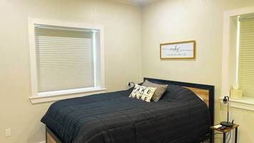 A bedroom with a bed, a bedside table with a lamp, a window with blinds, and a wall decoration.