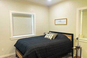 A bedroom with a bed, a bedside table with a lamp, a window with blinds, and a wall decoration.