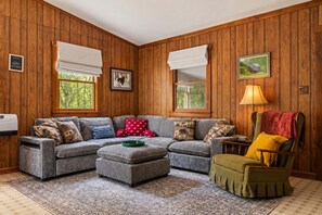 A cozy living room with a sectional sofa, ottoman, and a rocking chair. There are two windows with white blinds, a framed picture on the wall, and a lamp on a side table.