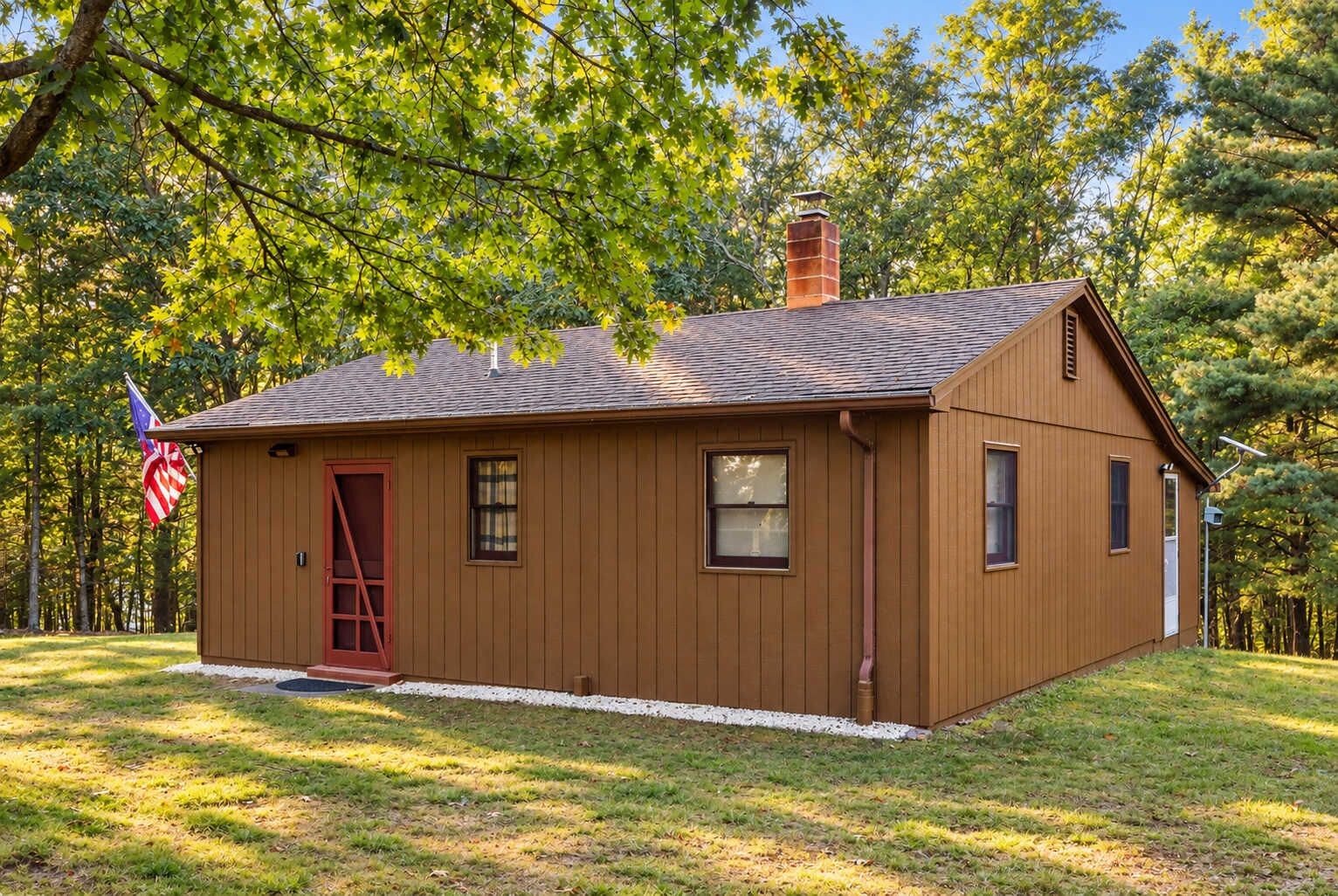 A small wooden cabin with a red door, windows, and a chimney, surrounded by trees and grass.