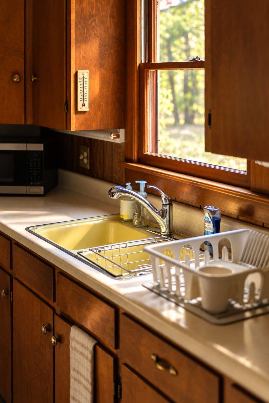 A kitchen with wooden cabinets, a stainless steel sink, a microwave, and a window with a view of trees.