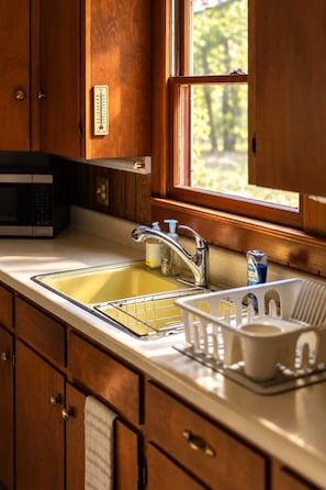 A kitchen with wooden cabinets, a stainless steel sink, a microwave, and a window with a view of trees.