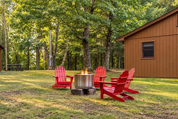 A wooden cabin with a fire pit and red Adirondack chairs in a wooded area.