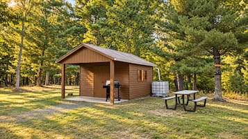 Un piccolo chalet in legno con veranda, barbecue e tavolo da picnic in una zona boschiva.