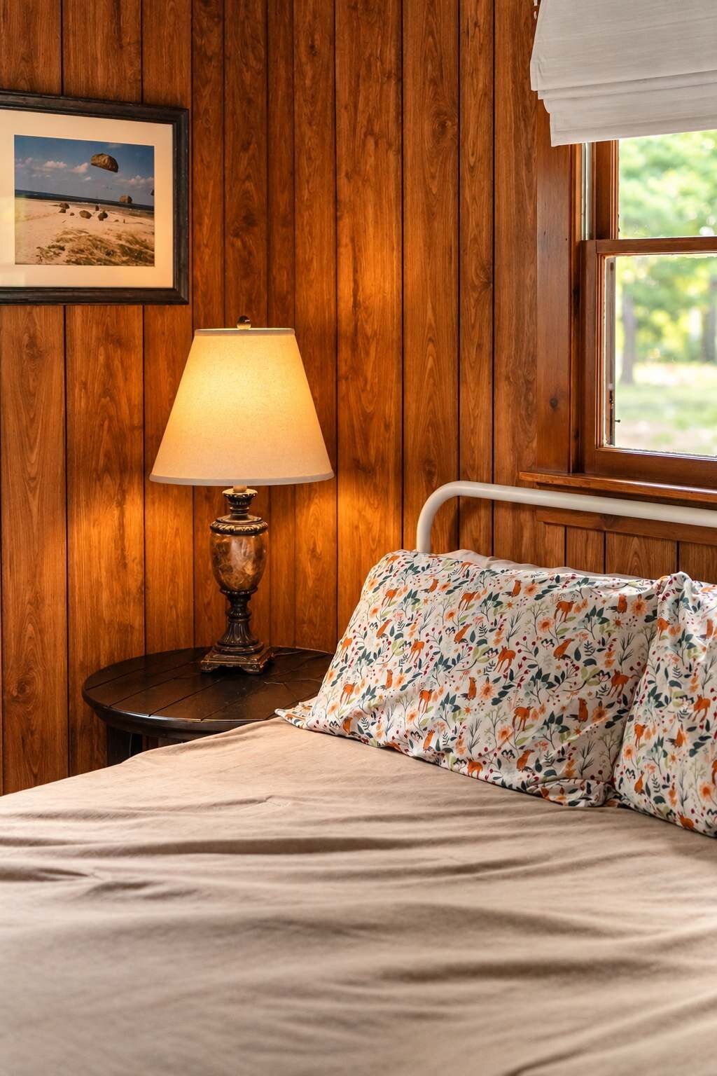 A bedroom with a wooden wall, a bedside lamp, a bed with floral bedding, and a window with a view of greenery.