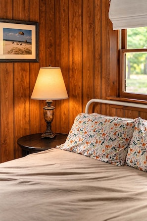 A bedroom with a wooden wall, a bedside lamp, a bed with floral bedding, and a window with a view of greenery.