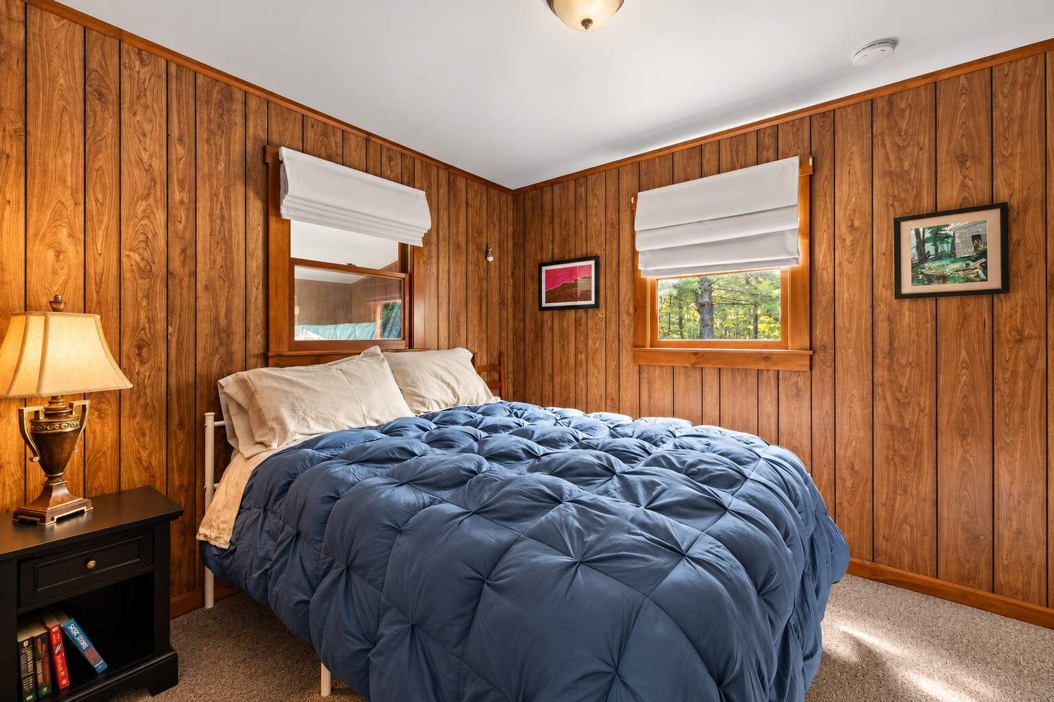 Ein Schlafzimmer mit Holzvertäfelung, einem Bett mit blauer Steppdecke, einem Nachttisch mit Lampe und einem Fenster mit Blick auf Bäume.
