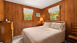 Un dormitorio con pared de paneles de madera, cama con ropa de cama floral, mesita de noche con lámpara y ventana con vistas a la vegetación.