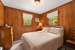Quarto com parede de madeira, cama com roupa de cama floral, mesa de cabeceira com abajur e janela com vista para o verde.