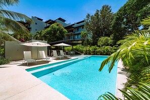 A swimming pool with lounge chairs and umbrellas, surrounded by lush greenery and modern buildings. - Chic Coastal Sanctuary 1-BR condo steps from the beach w/Private Terrace (Santa Cruz Huatulco)