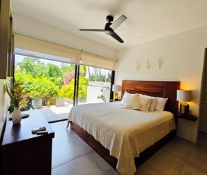 Ein Schlafzimmer mit einem großen Bett, einem Deckenventilator, einem Schreibtisch mit einer Vase und Gartenblick durch eine Schiebetür.