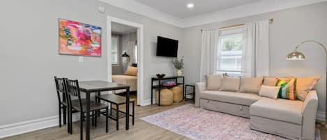 A living room with a sofa, a dining table with chairs, a TV mounted on the wall, and a window with curtains.
