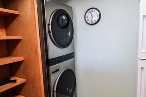 A laundry room with two stacked washers and dryers, wooden shelves, and a wall-mounted clock. - Beachview Condos #103 - Popular Family Retreat Just 30 Seconds from the Sand (South Padre Island)