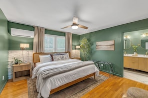 A bedroom with a bed, bedside tables, a nightstand, a ceiling fan, a window with blinds, a wall-mounted air conditioner, a mirror, and a bathroom vanity. - NEW - Maple Retreat - Hot Tub, Games, 8 min to NRG (Fayetteville)