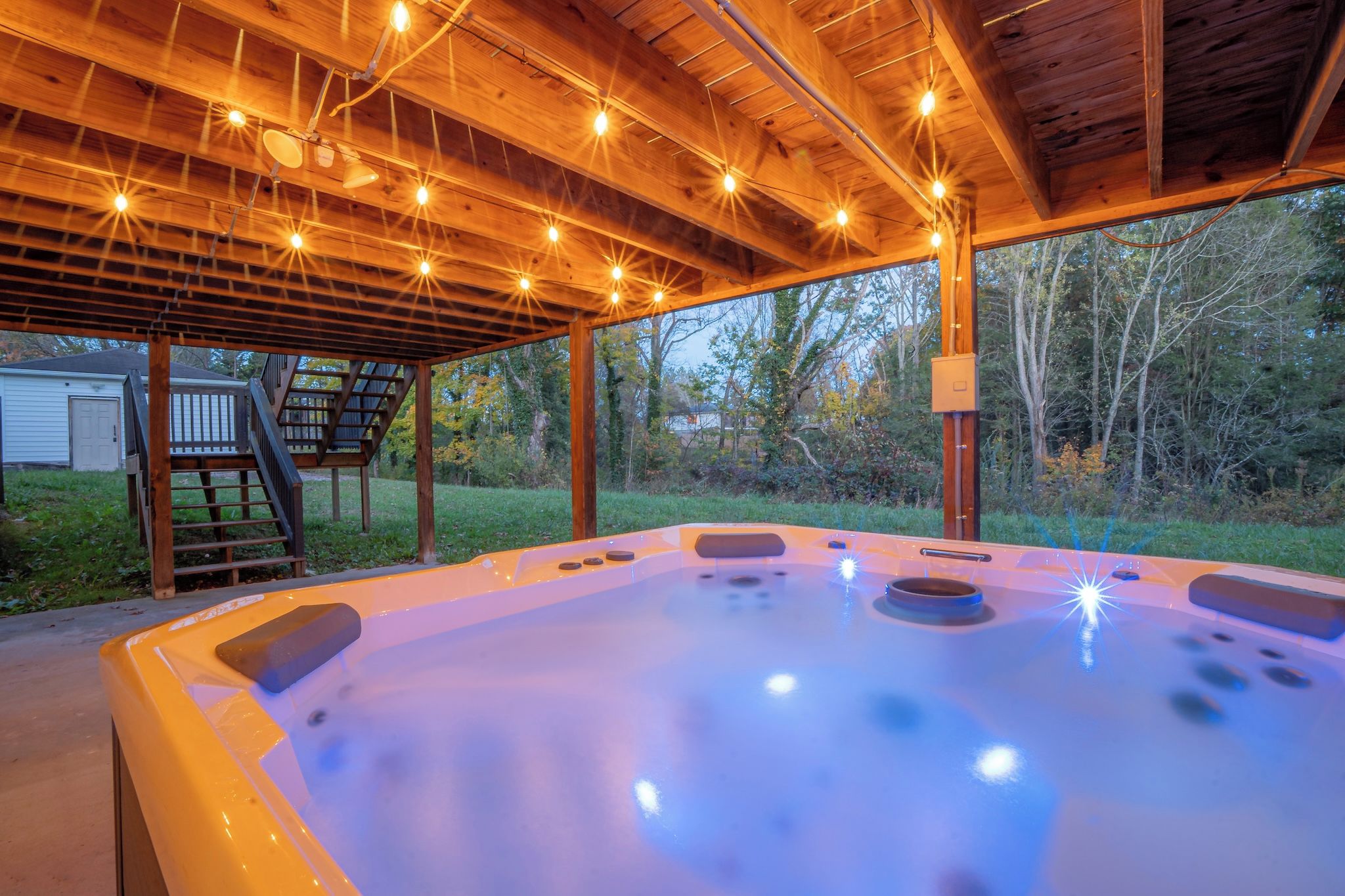 A hot tub under a wooden pergola with string lights, surrounded by a grassy yard and trees.