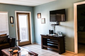 A living room with a television mounted on the wall, a coffee table with items, a door with a window, and a wall with framed pictures. - Beachview Condos #105 - "One of a Kind" Family Retreat Steps from the Gulf (South Padre Island)