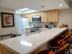 A modern kitchen with a marble countertop, wooden cabinets, a refrigerator, a microwave, a coffee maker, and a blender. - Beachview Condos #206 - Popular 2-BR Retreat with Pure Water & Netflix (South Padre Island)