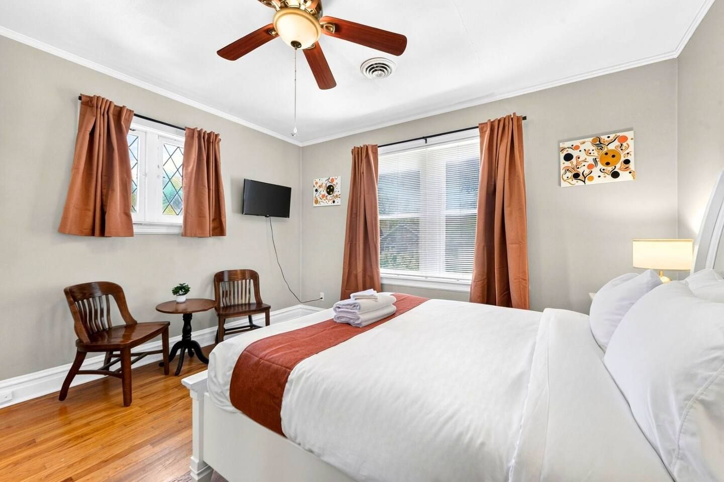Ein Hotelzimmer mit Bett, Schreibtisch, zwei Stühlen, Deckenventilator, Fernseher und einem Fenster mit Vorhängen.