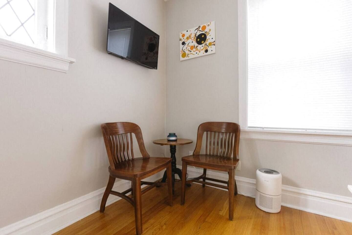 Ein Eckzimmer mit zwei Holzstühlen, einem kleinen runden Tisch, einem an der Wand befestigten Fernseher und einem Fenster mit Jalousien.