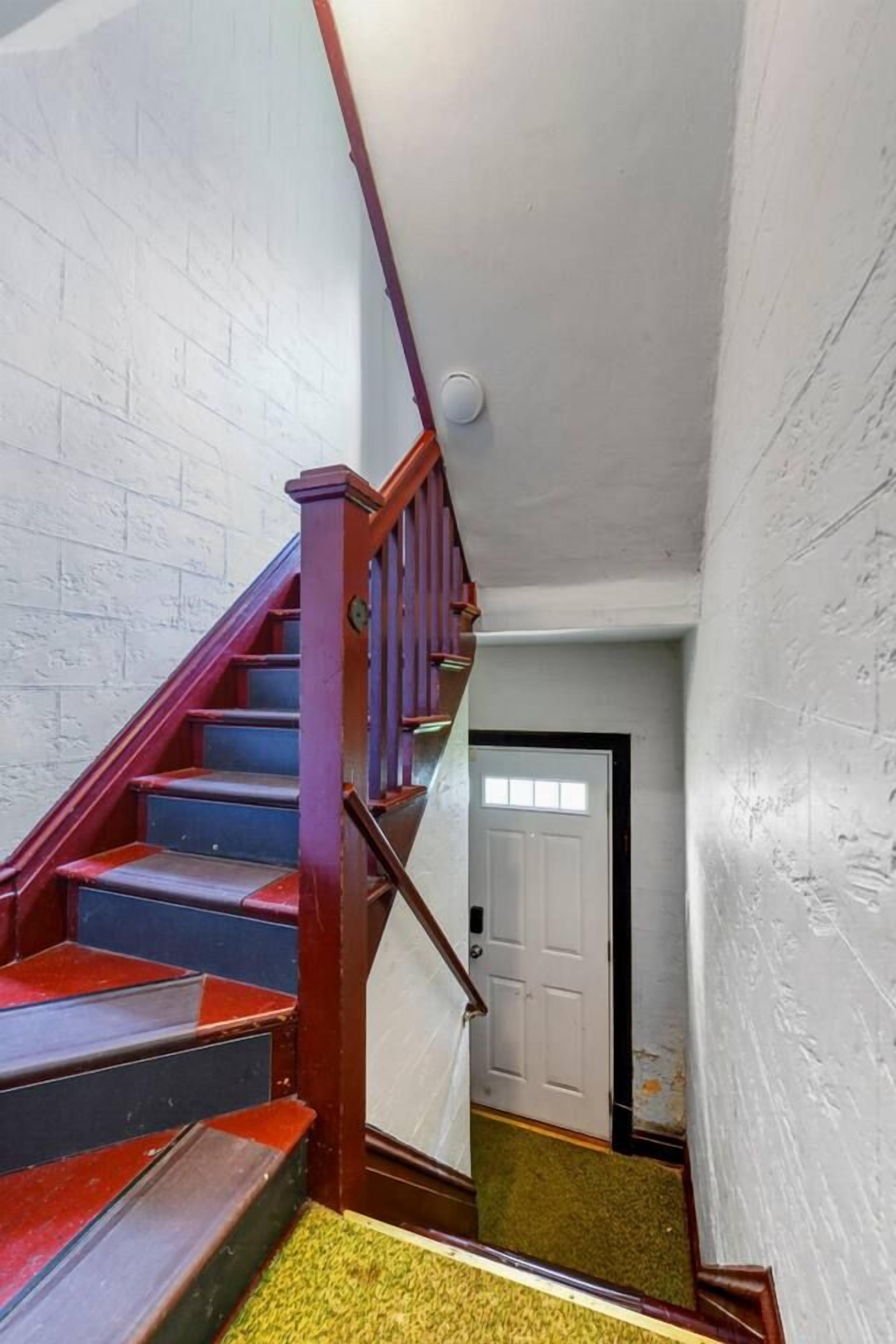 Una escalera con escalones rojos que conduce a una plataforma, una puerta blanca al final y una pared texturizada a la derecha.