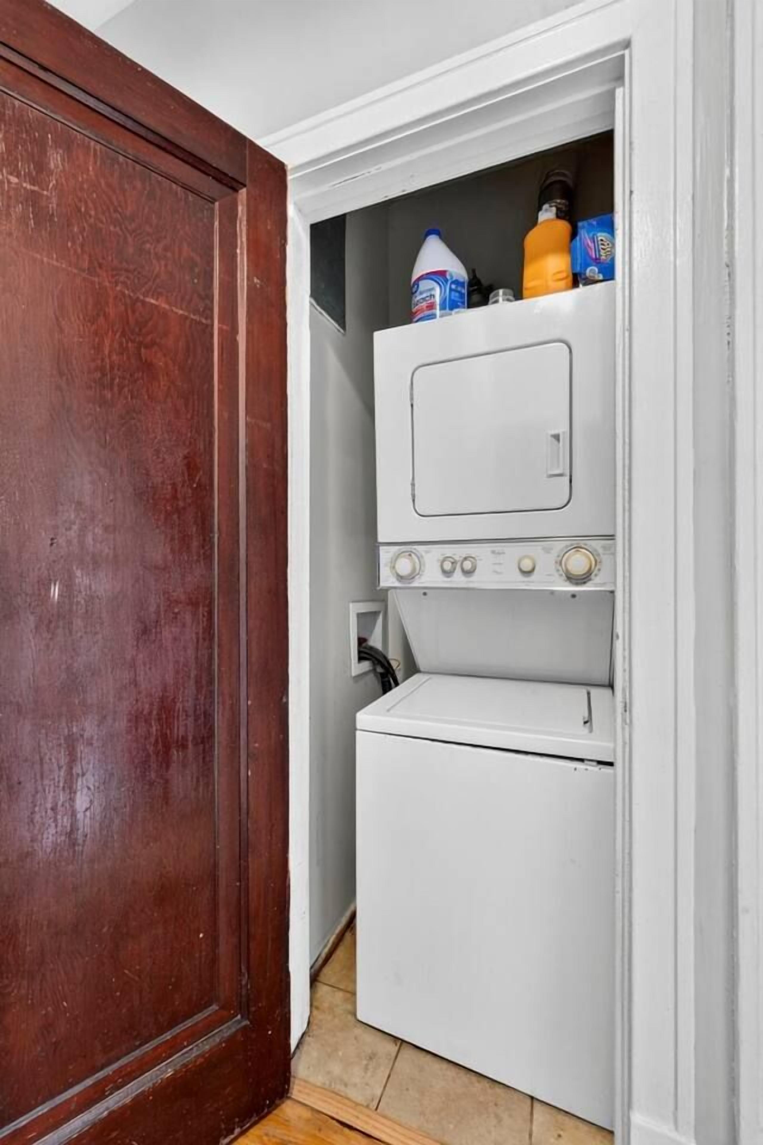 Un lavadero pequeño con lavadora y secadora blancas, una puerta de madera y un estante con productos de limpieza.