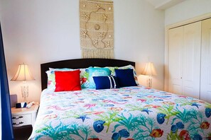 A bed with a colorful floral bedspread, two bedside lamps, a nightstand with a lamp, and a wall hanging above the bed. - Dolphin 28 - Spacious Families-Only Retreat Across from the Beach (South Padre Island)