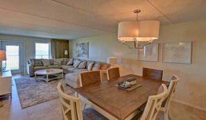 A living room with a sofa, coffee table, and dining table with chairs. There are two windows with curtains, a chandelier, and artwork on the walls. - Premier 3-BR Family Escape at Edgewater #507 - Spectacular Corner Gulf Views (South Padre Island)