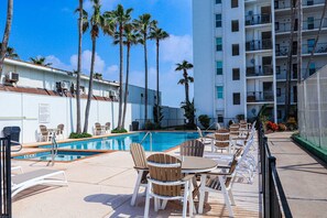 A pool area with lounge chairs and tables, surrounded by palm trees and a multi-story building. - Updated Beachfront Escape at Embassy #402 - Intimate 30-Unit Complex (South Padre Island)