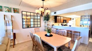A dining area with a round table, chairs, and a chandelier. There is a kitchen in the background with cabinets and appliances. - Updated Beachfront Escape at Embassy #402 - Intimate 30-Unit Complex (South Padre Island)