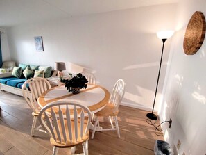 A modern living room with a round wooden dining table surrounded by white chairs, a sofa with patterned cushions, a floor lamp, and a wall-mounted light fixture. - Beachfront Value at Florence 1 #305 - Incredible Gulf Views (South Padre Island)